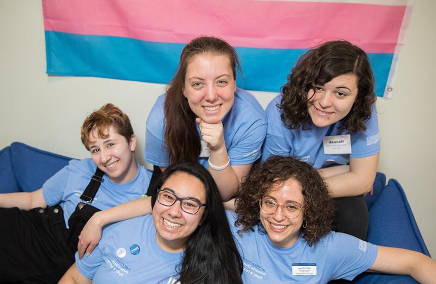 image of Current RWU students and staff in the Queer and Trans Resource and Advocacy Center in front of the trans pride flag